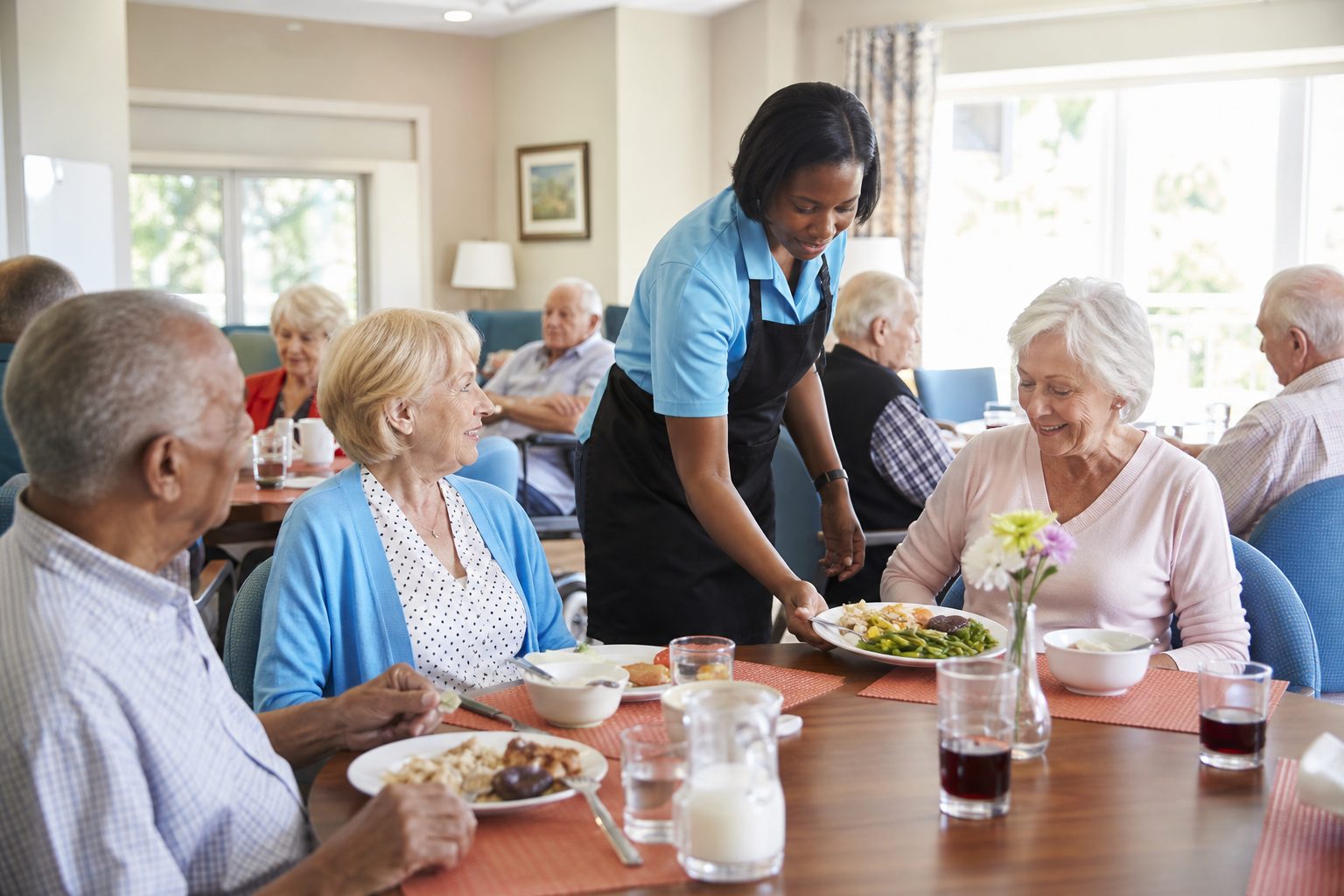Older adults seated at a senior day center dining table as a staff member serves the midday meal